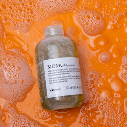 Bottle of shampoo labeled 'MOMO' on an orange background with soap bubbles