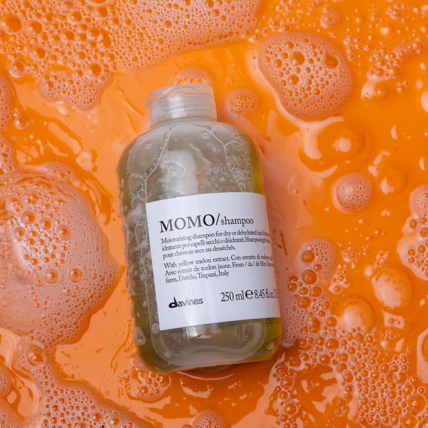 Bottle of shampoo labeled 'MOMO' on an orange background with soap bubbles