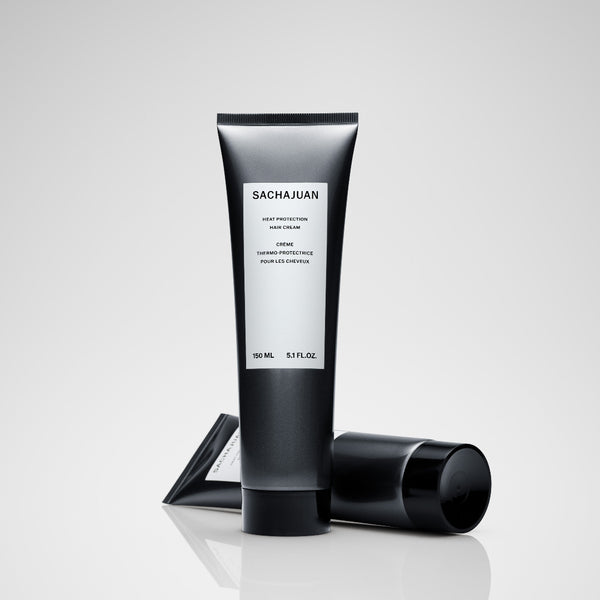 A black squeeze tube of Sachajuan Hair Cream with a white label, shown with its black cap lying beside it on a reflective light gray surface.