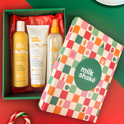 Milk Shake holiday gift set in a green box with red tissue paper, featuring a yellow shampoo bottle, a white body wash tube, and a clear hair oil bottle, alongside the branded gift box lid decorated with a colorful holiday checkered pattern.