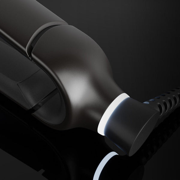 A close-up of a glossy black hair styling tool, likely a flat iron or curling iron, showing the heating plate and cord connector against a dark background.