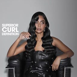 A woman seated in a black chair wearing a voluminous, long, black, defined curling hairstyle. The text 'SUPERIOR CURL DEFINITION' is printed in white on the left side against a dark gray gradient background.