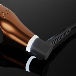 A close-up of a hair straightener with a metallic rose gold body and black accents, shown plugged in on a dark reflective surface.