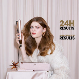 A woman with long wavy brown hair holds a pink and gold hair straightener in front of a light pink curtain backdrop. She is wearing a white faux fur coat, and a pink hair dryer sits on a table in the foreground.