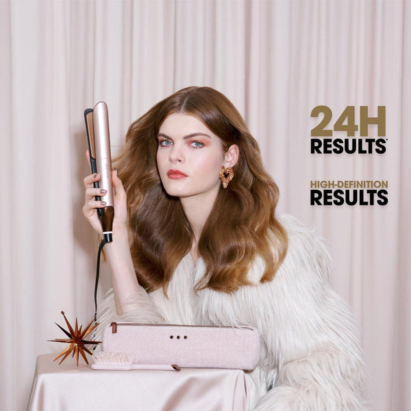 A woman with long wavy brown hair holds a pink and gold hair straightener in front of a light pink curtain backdrop. She is wearing a white faux fur coat, and a pink hair dryer sits on a table in the foreground.