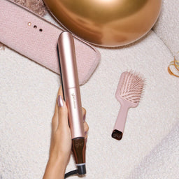 A hand holds a rose gold hair straightener on a white textured blanket next to a matching pink hairbrush, a pink pouch, and a gold ball.