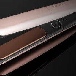 A close-up of a hair straightener shown open on a glossy black surface, with a rose gold exterior and a metallic bronze interior plate.