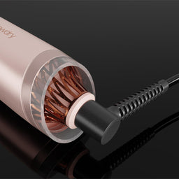 A close-up of a hair styling tool with a metallic rose gold body, showing the copper heating plates inside the barrel and a black cord plugged in on a glossy black background.