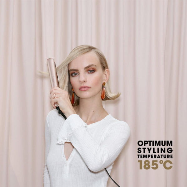 A woman with blonde hair holds a metallic rose gold flat iron against her hair, wearing a white long-sleeve top in front of a light pink curtain backdrop. Text on the right reads 'OPTIMUM STYLING TEMPERATURE 185°C'.