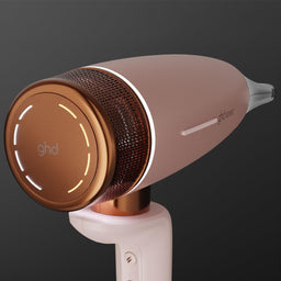 A GHD hair dryer in a rose gold finish with a metallic copper-toned circular front panel and illuminated LED accents, shown on a dark gray gradient background.