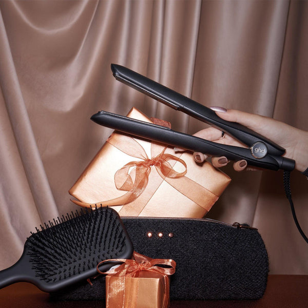 A hand holds a black hair straightener over a stack of gift boxes with metallic rose-gold and black packaging, with a black hairbrush in the foreground and a draped satin curtain backdrop.