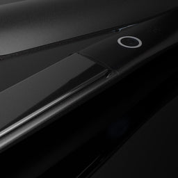 A close-up studio shot of a sleek black hair straightener, showing its smooth metallic plates and a small circular power button on a dark reflective background.