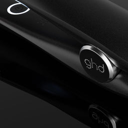 A close-up of a black ghd hair straightener with its power cord, showing the brand logo on the control dial against a dark reflective background.