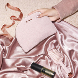 A hand holds a blush pink makeup pouch with three metal eyelets, styled on draped satin fabric with a pink hairbrush and a black-and-gold cosmetic tube nearby.