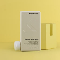Kevin Murphy Smooth.Again Wash smoothing shampoo in a tall off-white rectangular bottle, shown with its cap off beside it and a matching yellow box on a yellow background.