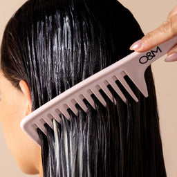 A close-up of a person with wet, dark hair being combed with a wide-tooth pink comb. A hand with manicured nails is visible holding the comb, set against a plain beige background.