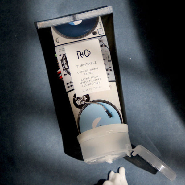 R+Co Turn Table Curl Defining Cream tube on a dark blue surface