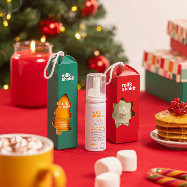Milk Shake whipped cream product displayed with its green and red gift boxes and a white pump bottle, set on a red tabletop with holiday decorations including a candle, wrapped gifts, marshmallows, and pancakes.