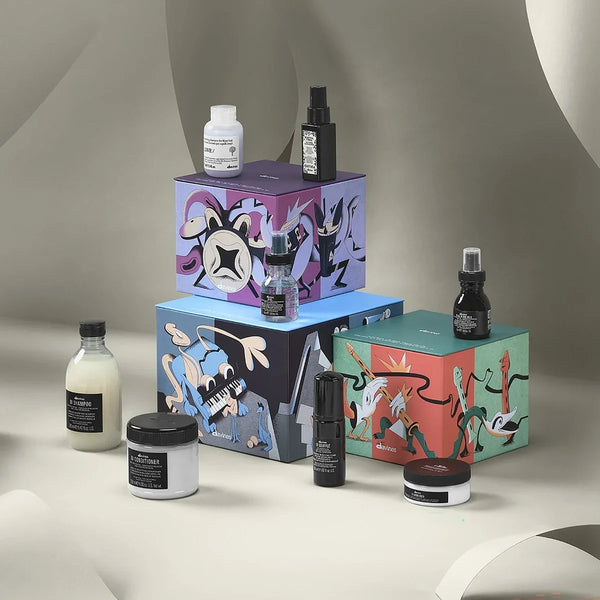 A curated beauty gift set featuring multiple cosmetic products and three colorful illustrated boxes, arranged on a neutral draped studio background. The products include jars, bottles, and a spray bottle with black labels, alongside purple, blue, and green packaging decorated with abstract line-art illustrations.