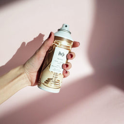 a can of R+Co Trophy Shine + Texture Spray held into the air with a pink background
